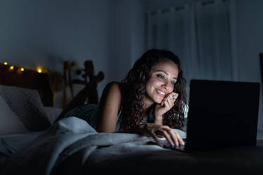 Vergnügte Frau in Nachtwäsche, die in einem dunklen Zimmer auf dem Bett liegt und sich ein lustiges Video auf einem Netbook ansieht, während sie lächelt und das Wochenende zu Hause verbringt - ADSF18766