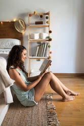 Side view of gentle female in pajamas sitting on floor near bed and enjoying interesting story while smiling and resting at home at weekend - ADSF18768