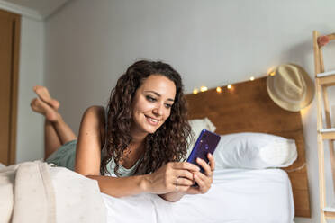 Low angle of carefree female lying on soft bed and reading messages on cellphone in morning after awakening - ADSF18770