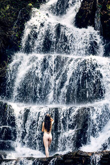 Rückenansicht nackte Frau mit langen Haaren stehen in der Nähe von Wasserfall und genießen die Natur - ADSF18773