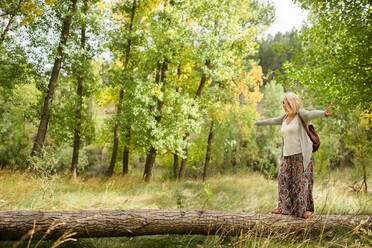 Full body of content blonde female traveler in stylish outfit balancing on fallen tree trunk with hands apart while enjoying summer day in green forest - ADSF18780