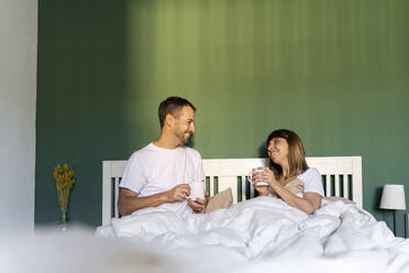 Smiling couple having coffee while sitting on bed at home - AFVF07713