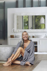 Happy mature woman looking away while sitting on floor by bathtub - MCF01506