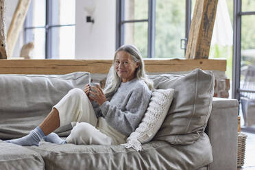 Lächelnde reife Frau hält eine Kaffeetasse, während sie zu Hause auf dem Sofa sitzt - MCF01513