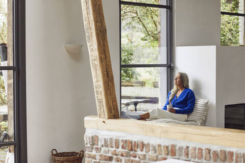 Nachdenkliche reife Frau mit Kaffeetasse, die durch ein Fenster zu Hause schaut, lizenzfreies Stockfoto