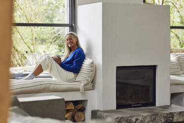 Smiling mature woman with coffee cup sitting on window seat at home - MCF01520