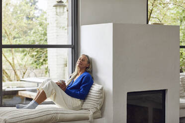 Mature woman with coffee cup day dreaming while leaning on wall of fireplace at home - MCF01521