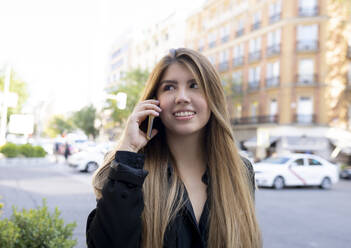 Lächelnde Frau, die auf der Straße in der Stadt steht und mit ihrem Handy telefoniert - JCCMF00092