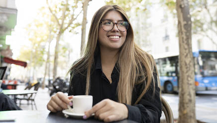 Young woman with coffee cup smiling while sitting at sidewalk cafe in city - JCCMF00093