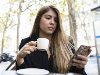 Frau, die ein Mobiltelefon benutzt, während sie in einem Straßencafé in der Stadt Kaffee trinkt - JCCMF00095