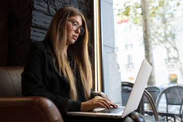 Junge Frau mit Brille, die einen Laptop benutzt, während sie in einem Café sitzt - JCCMF00098