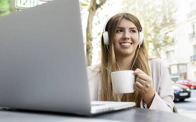 Frau mit Kopfhörern trinkt Kaffee, während sie mit einem Laptop in einem Straßencafé sitzt - JCCMF00113