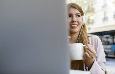 Smiling young woman with laptop and coffee cup looking away while sitting at sidewalk cafe - JCCMF00114