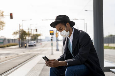 Young man using mobile phone at bus stop during COVID-19 - AFVF07722