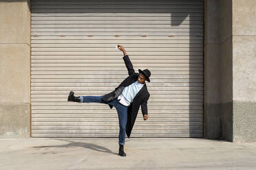 Modischer junger Mann steht auf einem Bein und tanzt gegen einen Fensterladen - AFVF07755