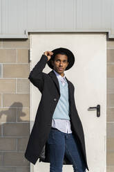 Modischer junger Mann mit Hut, der an einem sonnigen Tag vor einer Tür steht - AFVF07764
