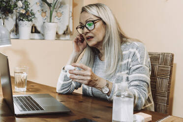 Senior woman wearing wireless in-ear headphones during online consultation at home - ERRF04795