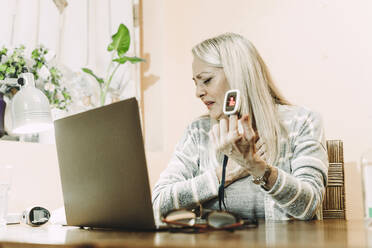 Ältere Frau in Angst zeigt Pulsoximeter auf Laptop während Online-Beratung zu Hause - ERRF04808