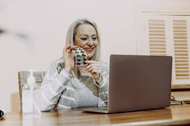 Lächelnde ältere Frau, die während einer Online-Konsultation zu Hause Medikamente zeigt - ERRF04817