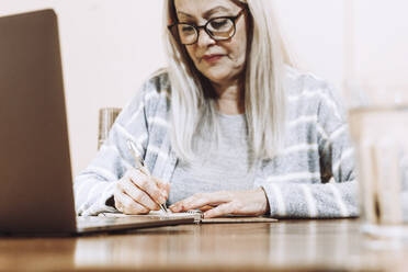 Ältere Frau schreibt Notizen während einer Online-Konsultation zu Hause - ERRF04818