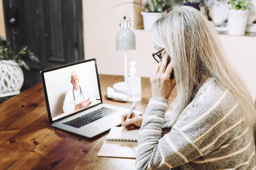 Ältere Frau, die mit einem Mobiltelefon spricht, während sie bei einem Videogespräch mit einem Arzt zu Hause Notizen schreibt - ERRF04819