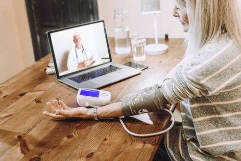 Ältere Frau prüft ihren Blutdruck unter Anleitung eines Arztes per Videoanruf zu Hause - ERRF04824