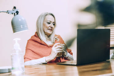 Ältere Frau hält ein Trinkglas während einer Online-Beratung zu Hause - ERRF04832