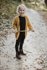 Lächelndes Mädchen, das mit einem Stock spielt, während es auf einem Fußweg im Wald steht - GMLF00879