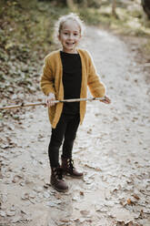 Smiling girl playing with stick while standing on footpath in forest - GMLF00879