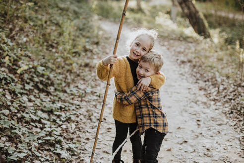 Schwester hält Stock und umarmt Bruder im Wald stehend - GMLF00880