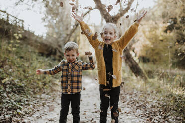 Happy sibling playing with dry fallen leaf while standing in forest during autumn - GMLF00881