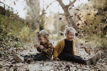 Cheerful girl and boy playing with dry fallen leaf while sitting on footpath in forest - GMLF00883