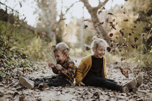 Fröhliches Mädchen und Junge spielen mit trockenen, gefallenen Blättern, während sie auf einem Fußweg im Wald sitzen - GMLF00883