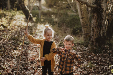 Girl and boy playing with stick and rope while standing in forest during autumn - GMLF00884