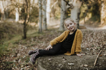 Smiling girl relaxing on concrete bench in forest during autumn - GMLF00896