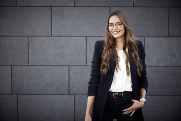 Smiling female entrepreneur standing with hands in pockets against brick wall - RHF02462