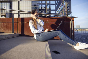 Glückliche Geschäftsfrau arbeitet am Laptop, während sie bei Sonnenuntergang auf den Stufen sitzt - RHF02491