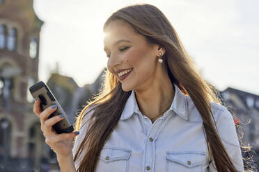 Happy businesswoman using mobile phone while standing in city - RHF02517