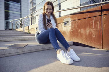 Businesswoman using mobile phone while sitting by laptop on steps during sunset - RHF02523