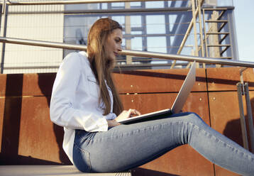 Junge Geschäftsfrau, die auf einer Treppe sitzend am Laptop arbeitet - RHF02524