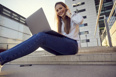 Lächelnde Geschäftsfrau mit Hand im Haar, die einen Laptop benutzt, während sie auf einer Treppe sitzt - RHF02525