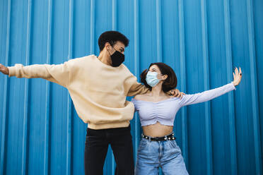 Unbekümmertes Paar mit Gesichtsmaske tanzt an einer blauen Wand - MIMFF00283