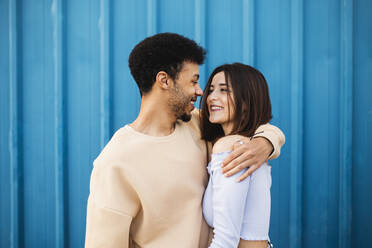 Smiling young man embracing woman while standing against blue wall - MIMFF00292