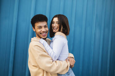 Glückliches junges Paar, das sich gegenseitig umarmt, während es an einer blauen Wand steht - MIMFF00293