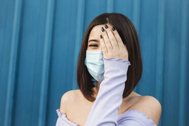 Young woman wearing face mask covering eye with hand while standing against blue wall - MIMFF00295