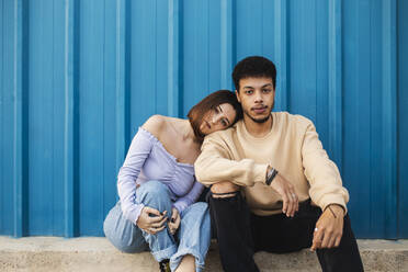 Girlfriend leaning on boyfriend's shoulder while sitting against blue wall - MIMFF00296