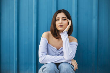 Young beautiful woman sitting against blue wall - MIMFF00297