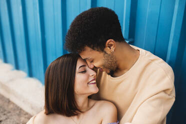 Lächelndes junges Paar sitzt von Angesicht zu Angesicht an einer blauen Wand - MIMFF00298