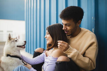 Smiling couple sitting with dog while leaning on blue wall - MIMFF00299