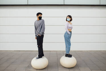 Couple with face mask looking over shoulder while standing on white concrete ball - MIMFF00301